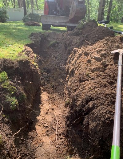 A freshly dug trench in a grassy yard with a shovel and construction equipment visible in the background.