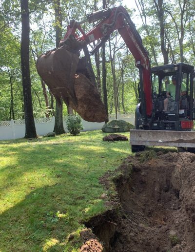 A person operates an excavator to lift a large rock out of a hole in a grassy, tree-lined backyard.