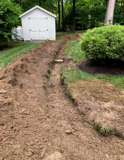 A narrow dirt trench runs through a grassy yard toward a white shed, with displaced soil and removed grass visible beside the trench.
