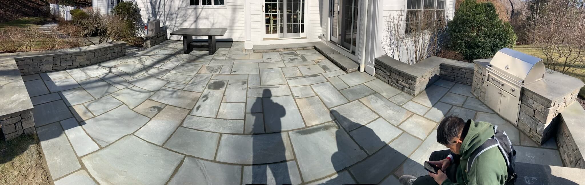 Panoramic view of a stone patio with outdoor furniture, retaining walls, and a person's shadow visible on the ground.